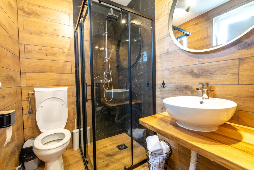 Baño reformado con mampara negra, lavabo sobre encimera de madera y paredes revestidas de madera