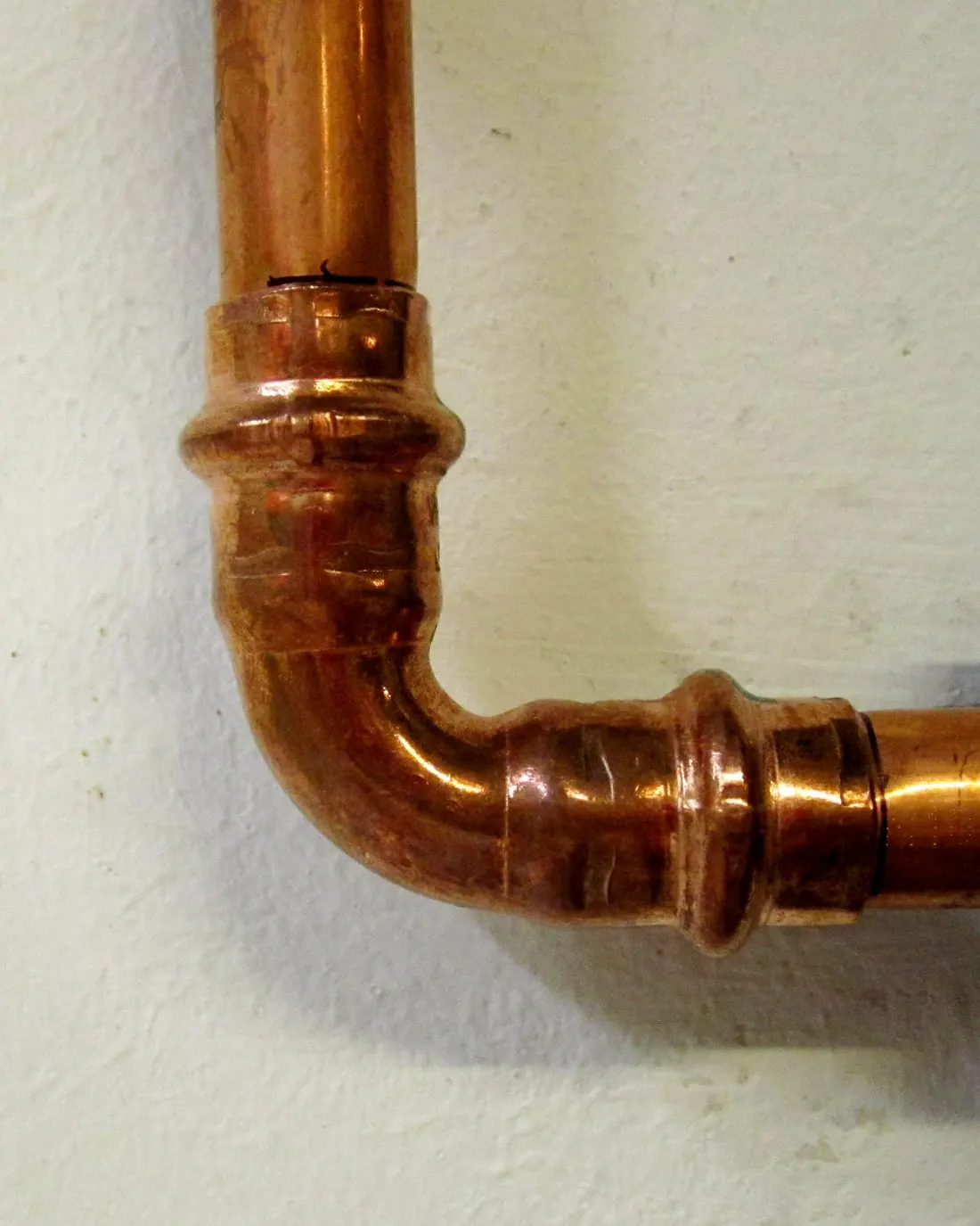 Codo de cobre prensado uniendo dos tramos de tubería junto a una pared blanca